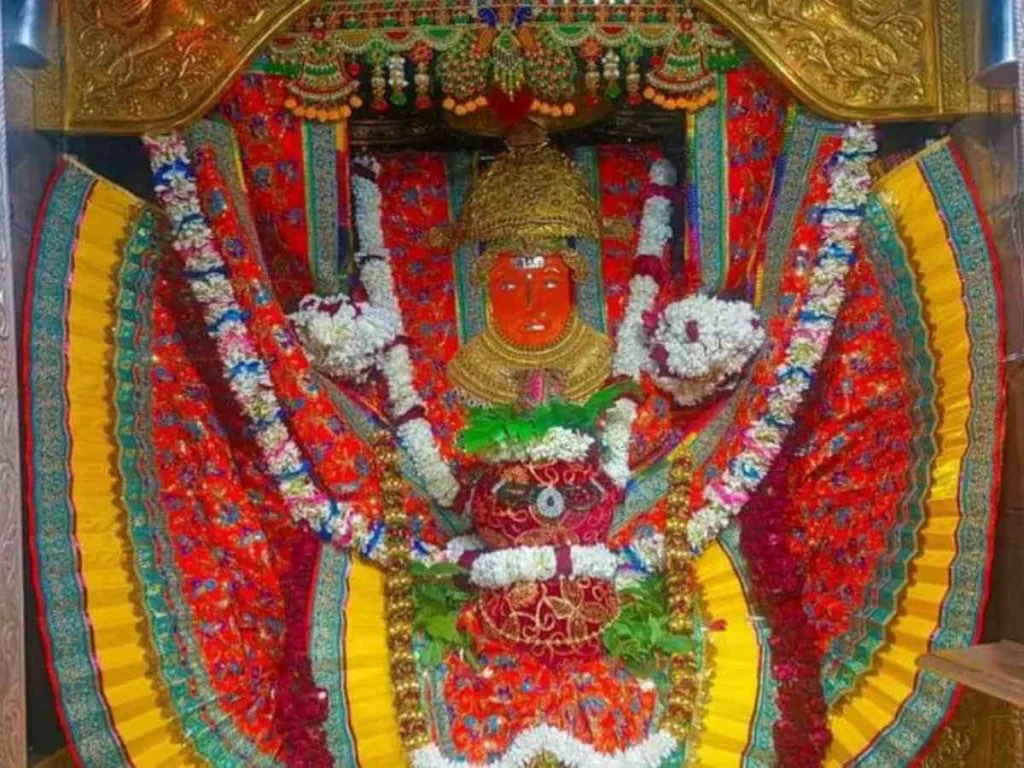 Full darshan of Jeen Mata in the sanctum with golden mukut, sacred kalash, white and red garlands and festive red, yellow and green drapery under a gilded arch