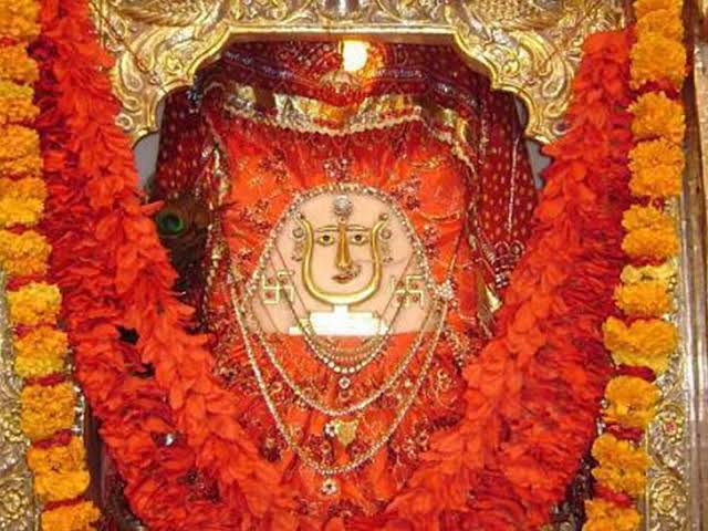 Ancient silver mukhota face of Jeen Mata with swastika symbols, framed by orange and yellow marigold garlands in a gilded shrine